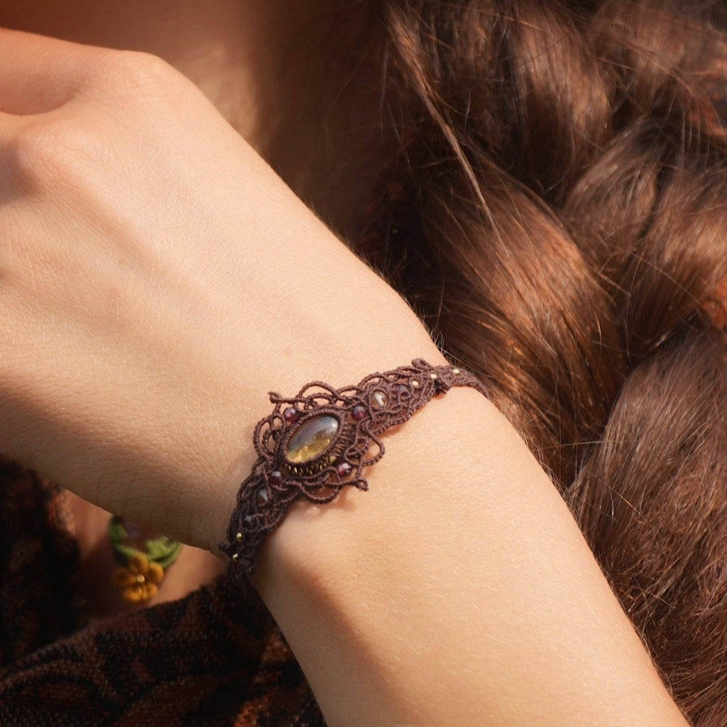 Bracelet large elfique en micro macramé brun, orné d’une citrine jaune pâle et de perles de grenat rouge rosé. Bijou bohème et artisanal réglable, porté au poignet d’une femme à la peau claire.