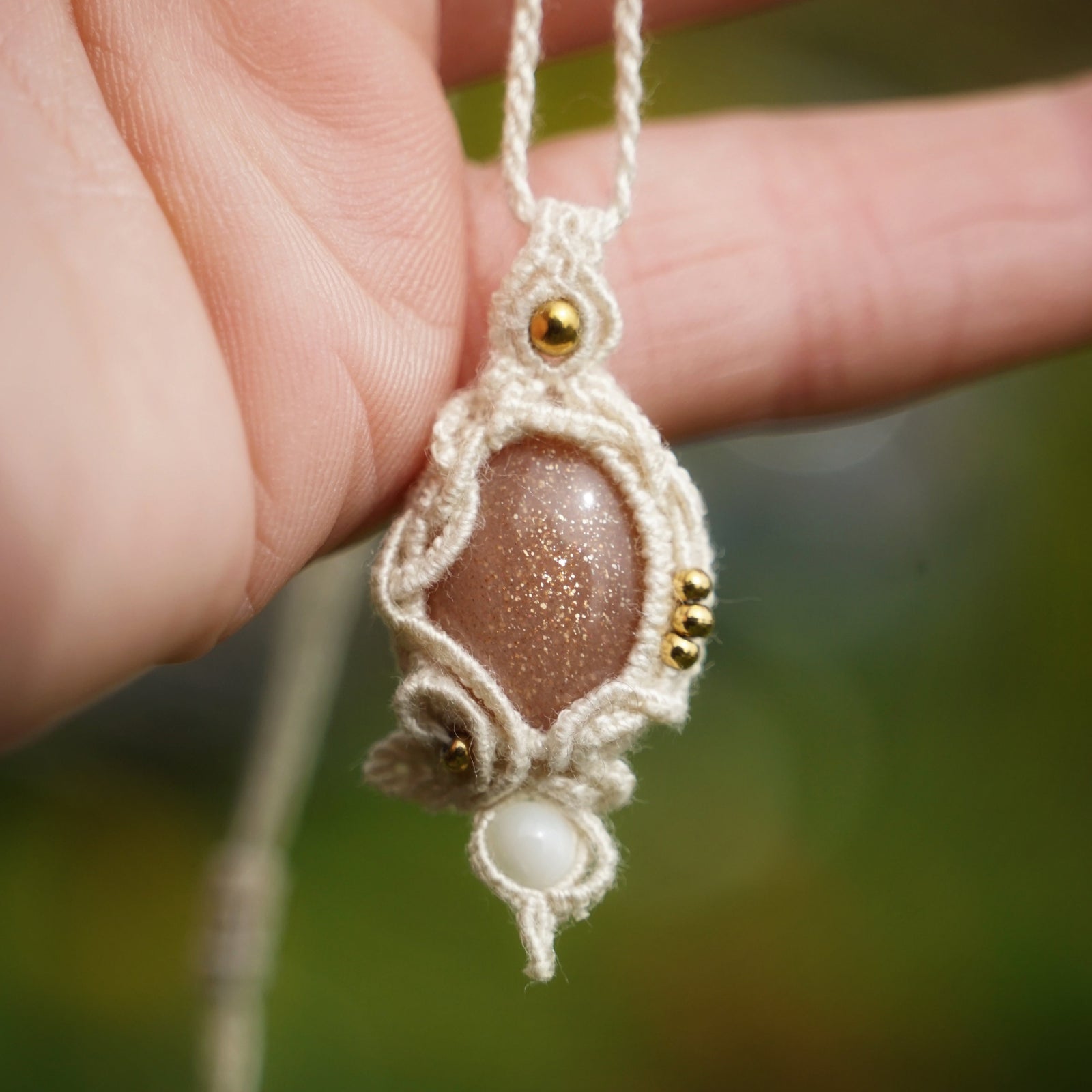 Collier bohème en micro macramé coton écru avec pierre de soleil pêche et perles de nacre, tenu dans une main délicate, fond vert naturel. Coton et Astrance