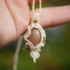 Collier bohème en micro macramé coton écru avec pierre de soleil pêche et perles de nacre, tenu dans une main délicate, fond vert naturel. Coton et Astrance