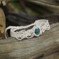 Bracelet large asymétrique. Bijou dentelle ambiance bohème hippie. Bracelet tissé en micro macramé avec fil de coton écru de 0.5 mm avec une pierre ovale de chrysocolle couleur bleu turquoise et marron foncé chocolat. Présence de part et d'autre de façon symétrique des perles en acier inoxydable argentées. Système de réglage à coulisse tubulaire à l'arrière. Design en relief et en torsade entrelacées celtiques. Ambiance plages, festival d'été et mariage boho. Bijou posé sur un galet beige et coquillages.