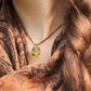 Collier avec pendentif en micro macramé avec motifs floraux. Bijou féminin composé d'un citrine jaune pâle translucide ovale sertie dans une coque marron foncé tissé avec un fil de 0.5 mm sur lequel est fixé une liane verte entrelacée avec mini feuilles et une fleur à 5 pétales jaune orangé avec un cœur doré de 2 mm.

Le bijou est porté autour d'un cou de femme à la peau claire et d'un châle orange et noir et natte brune. le bijou est pris de face avec rayon du soleil
