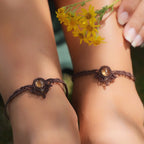 Bijoux de pieds, paire assortie de bracelets de chevilles à ambiance bohème celtique elfique. Fil marron foncé fin tissé en micro macramé avec entrelacs et arabesques, pierre centrale ovale petite taille jaune miel translucide. Bijoux hippie, bohème avec inspiration nature. Chevillères portées sur des chevilles à la peau pâle, bijoux de face avec jeu d'ombre et lumière et fleurs jaune en haut à droite.