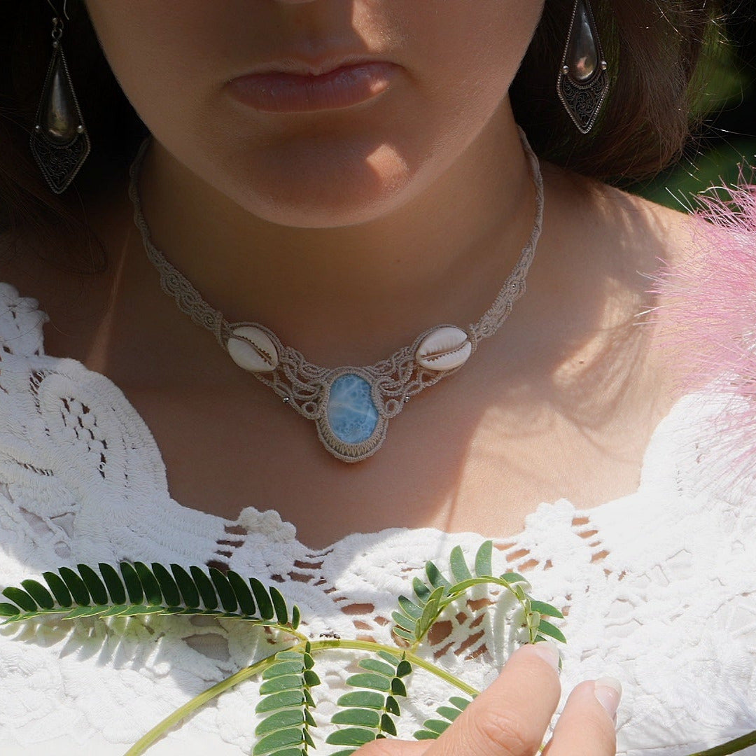 Collier plastron en Larimar Océan bleu ciel, coton couleur lin naturel (beige gris), cauris blancs et perles en acier inoxydable argenté. Système de réglable en tube tissé à l'arrière du bijou. Tissage symétrique en micro macramé avec effet nœuds celtiques entrelacés en relief. Style bohème, hippie, plage, Sirène, tribal et tropical. Convient pour un mariage type bohème ou plage. Bijou porté avec une tenue romantique et bohème blanche avec dentelle apparente. Modèle à la peau bronzée et aux cheveux bruns. 