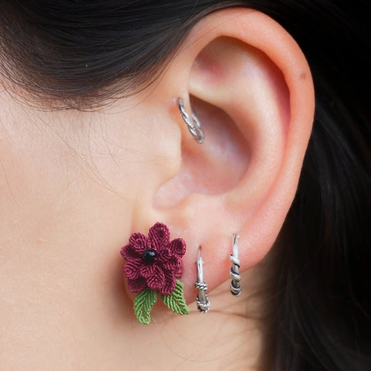 Boucles d’oreilles florales en micro macramé faites main, collection « Pétales éternels ». Bijou artisanal pour femme en coton rouge bordeaux et vert tendre, tissé en fleur 3D avec 13 pétales, 2 feuilles et un cœur en perle d’onyx noire de 3 mm. Fixées sur clous d’oreilles hypoallergéniques en acier inoxydable argenté avec poussoirs papillons. Style bohème, elfique et hippie, idéal pour festivals et costumes féériques. Photo portée sur modèle femme à peau claire et cheveux bruns.