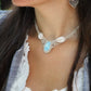 Collier plastron en Larimar Océan bleu ciel, coton couleur lin naturel (beige gris), cauris blancs et perles en acier inoxydable argenté. Système de réglable en tube tissé à l'arrière du bijou. Tissage symétrique en micro macramé avec effet nœuds celtiques entrelacés en relief. Style bohème, hippie, plage, Sirène, tribal et tropical. Convient pour un mariage type bohème ou plage. Bijou porté avec une tenue marine bohème bleu et blanc avec dentelle apparente. Modèle à la peau bronzée et aux cheveux bruns. 