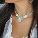 Collier plastron en Larimar Océan bleu ciel, coton couleur lin naturel (beige gris), cauris blancs et perles en acier inoxydable argenté. Système de réglable en tube tissé à l'arrière du bijou. Tissage symétrique en micro macramé avec effet nœuds celtiques entrelacés en relief. Style bohème, hippie, plage, Sirène, tribal et tropical. Convient pour un mariage type bohème ou plage. Bijou porté avec une tenue marine bohème bleu et blanc avec dentelle apparente. Modèle à la peau bronzée et aux cheveux bruns. 