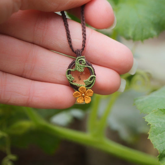 Collier avec pendentif en micro macramé avec motifs floraux. Bijou féminin composé d'un citrine jaune pâle translucide ovale sertie dans une coque marron foncé tissé avec un fil de 0.5 mm sur lequel est fixé une liane verte entrelacée avec mini feuilles et une fleur à 5 pétales jaune orangé avec un cœur doré de 2 mm.

Le bijou est en verticale avec main à l'arrière. Le bijou est pris de face avec une certaine transparence et un fond vert naturel, au milieu des tortillons et feuilles de courges