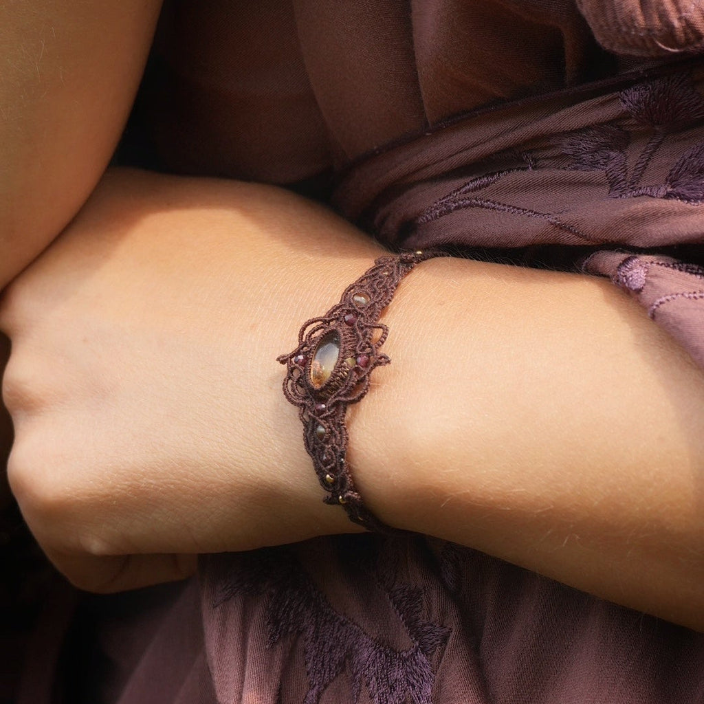 Bracelet large elfique en micro macramé brun, orné d’une citrine jaune pâle et de perles de grenat rouge rosé. Bijou bohème et artisanal réglable, porté au poignet d’une femme à la peau claire.