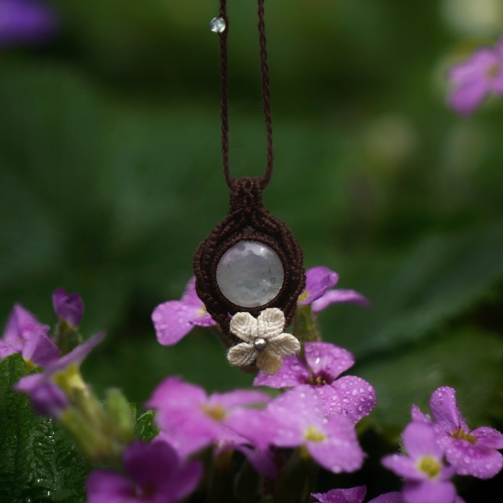 Bijou pendentif en labradorite blanche suspendu verticalement au milieu de fleurs roses et de feuilles de fraisiers mouillées par la pluie, collier elfique bohème réglable. Coton et Astrance