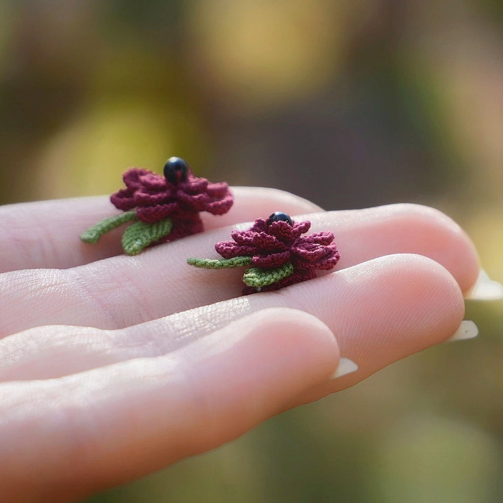 Boucles d’oreilles florales en micro macramé faites main, collection « Pétales éternels », posées sur une main féminine. Fleur 3D tissée en coton rouge bordeaux et vert tendre avec 13 pétales, 2 feuilles et cœur en perle d’onyx noire. Style bohème, elfique et hippie, idéal pour festivals et costumes féériques, photographié au milieu de feuilles et de nature verdoyante.