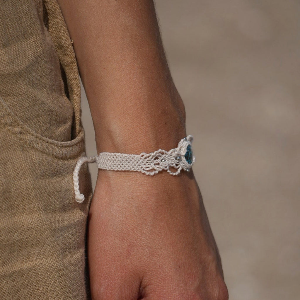 Bracelet large asymétrique. Bijou dentelle ambiance bohème hippie. Bracelet tissé en micro macramé avec fil de coton écru de 0.5 mm avec une pierre ovale de chrysocolle couleur bleu turquoise et marron foncé chocolat. Présence de part et d'autre de façon symétrique des perles en acier inoxydable argentées. Système de réglage à coulisse tubulaire à l'arrière. Design en relief et en torsade entrelacées celtiques. Ambiance plages, festival d'été, tribal. Bijou porté fond mer et ciel, pantalon beige lagre.